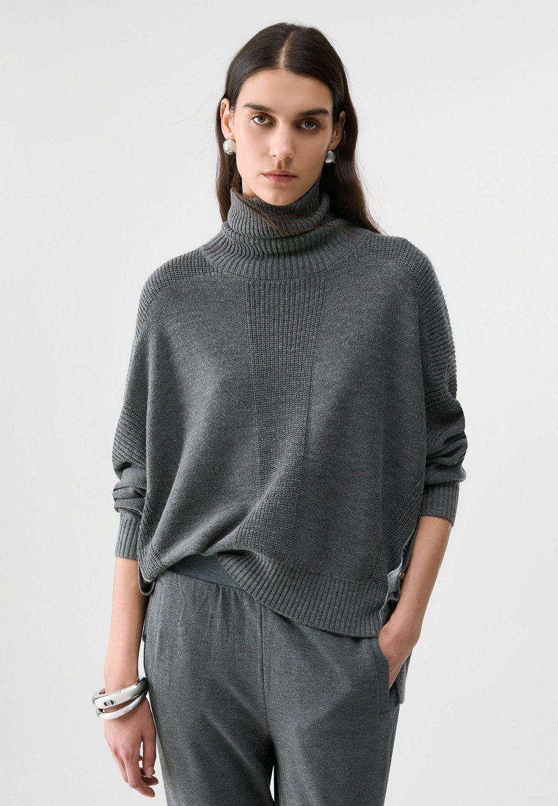 Femme aux longs cheveux foncés porte un pull en maille gris à col montant et un pantalon gris assorti, accessoirisés de larges boucles d’oreilles et bracelets argentés.