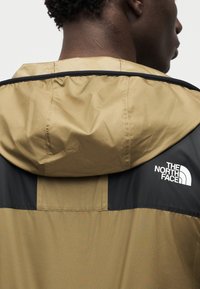 Αδιάβροχο μπουφάν χακί και μαύρο με κουκούλα, με έντονο λευκό λογότυπο στον δεξιό ώμο και λεία, ελαφριά υφή υφάσματος.