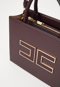 Sac à main en cuir violet avec quincaillerie dorée, de forme rectangulaire, logo embossé et fermeture à glissière. Texture lisse et design structuré.