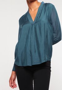 Blusa teal con scollo a V, maniche lunghe e vestibilità comoda; realizzata in un tessuto morbido e lucido; presenta una trama sottile e un orlo arrotondato.
