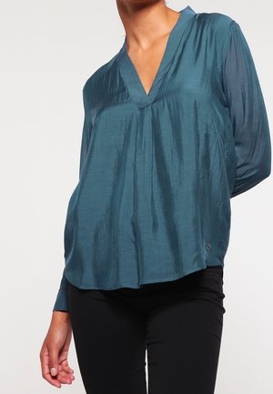 Blouse teal avec un décolleté en V, des manches longues et une coupe décontractée ; fabriquée à partir d'un tissu lisse et brillant ; dispose d'une texture subtile et d'un ourlet arrondi.