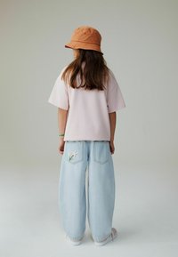 Maglietta rosa a maniche corte, jeans a gamba larga azzurri con fiori ricamati sulle tasche posteriori e cappello bucket arancione.