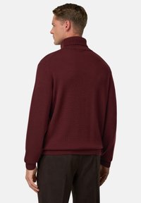 Pull en maille bordeaux avec un col montant, des poignets et un ourlet côtelés ; présente une coupe décontractée et un tissu texturé. Le modèle se tient de dos.