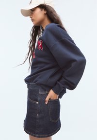 Ung kvinna iklädd vit keps, marinblå sweatshirt med röd text och mörk denimkjol, poserar med handen i fickan mot en enkel bakgrund.