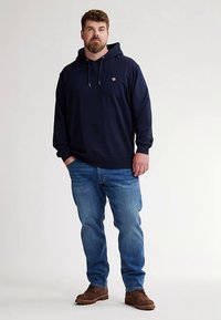 Hanorac din bumbac albastru marin cu buzunar frontal și șnururi, asortat cu blugi deschis la culoare și cizme maro, având un mic logo pe piept.