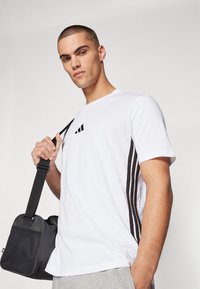 Witte korte mouwen t-shirt met zwart drie-strepens ontwerp en logo detail. Het model houdt een compacte zwarte schoudertas vast met een gestructureerd oppervlak.