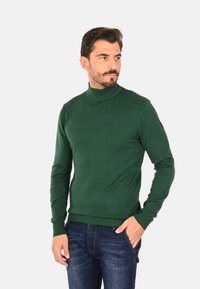 Maglione verde a collo alto in maglia, polsini e orlo a coste, design aderente, abbinato a jeans di denim blu.