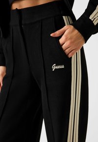 Schwarze Jogginghose mit strukturierter Oberfläche, zwei Fronttaschen, Falten und cremefarbenen Streifen an den Seiten. "Guess" in Weiß gestickt.