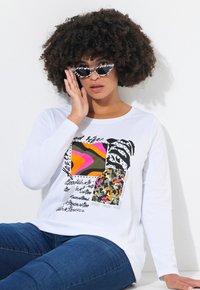 Weißes Langarm-T-Shirt mit einem bunten grafischen Design in Pink-, Grün- und Schwarzmustern. Kombiniert mit schwarz-weißen Katzenaugen-Sonnenbrillen.