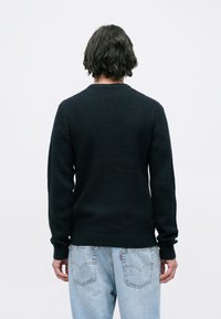 Άτομο με αφέλειες και σκούρα μαλλιά στο ύψος των ώμων φοράει ένα μαύρο πλεκτό πουλόβερ και ανοιχτό μπλε τζιν, στέκεται γυρισμένο προς τα πίσω σε έναν απλό φόντο.