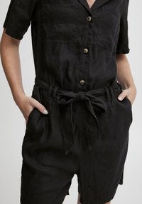 Juoda lininė šokinė su trumpomis rankovėmis, sagčių priekyje, dviem krūtinės kišenėmis ir dirželiu juosmenyje. Siūlo laisvą prigludimą ir švelnią tekstūrą.