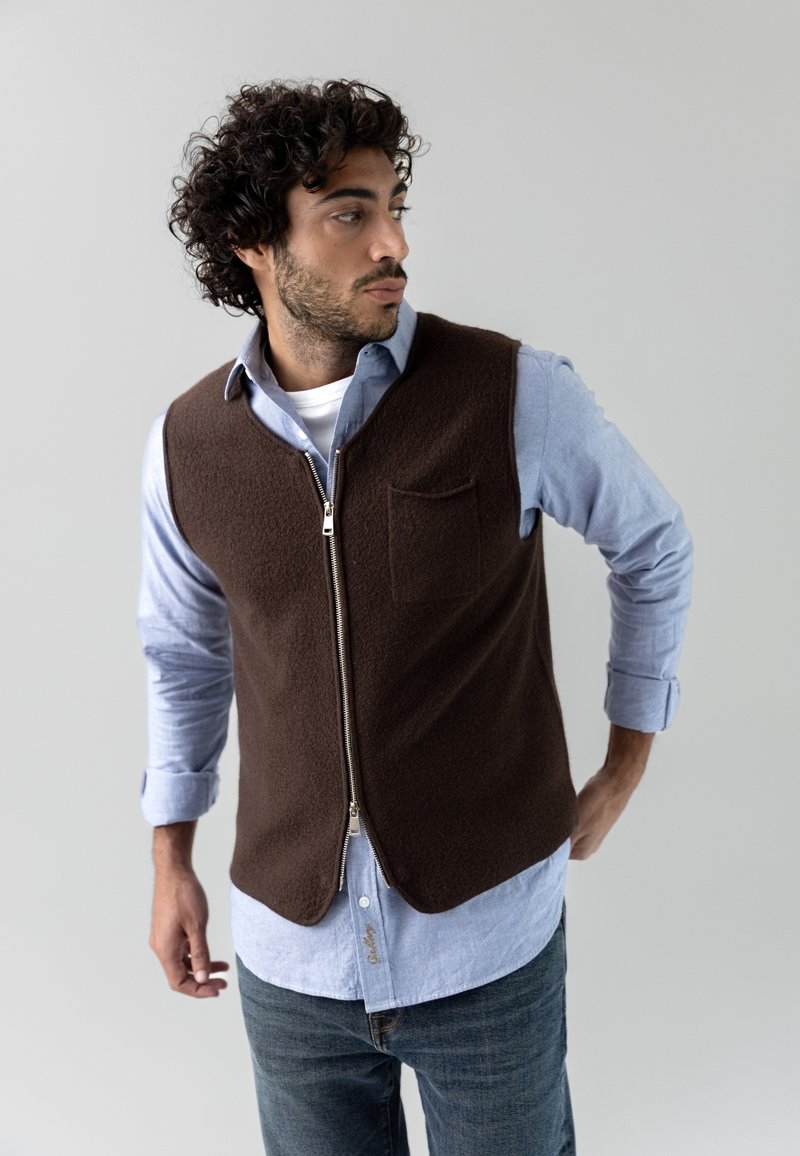Gilet in pile marrone con zip frontale, dotato di tasca sul petto. Indossato sopra una camicia azzurro chiaro con bottoni, abbinato a jeans denim casual.