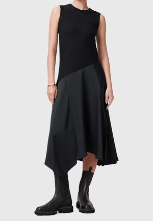 Vrouw in een mouwloze zwarte jurk met asymmetrische zoom en zwarte grove laarzen tot halverwege de kuit, staand tegen een egale lichte achtergrond.