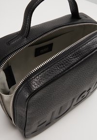 Schwarze Ledertasche mit strukturierten Oberflächen, ausgestattet mit einem silbernen Reißverschluss und einem beigen Innenleben. "HUGO"-Logo auf einer Seite eingeprägt.
