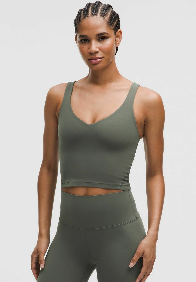 Donna con capelli intrecciati che indossa un top sportivo verde oliva e leggings a vita alta, in piedi contro uno sfondo chiaro uniforme.