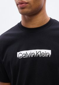 Μαύρο βαμβακερό μπλουζάκι με εμφανές λευκό λογότυπο Calvin Klein στο στήθος. Η λαιμόκοψη είναι κλασικό στρογγυλό λαιμό.