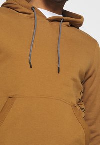 Hoodie marron en tissu doux avec une poche kangourou, des cordons noirs et des détails de couture latérale. Comprend un design graphique discret.