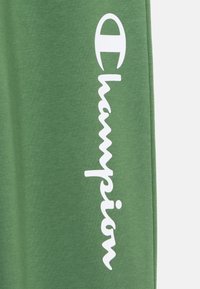 Zaļi kokvilnas svārki ar baltu "Champion" logo slīprakstā gar sānu. Audums izskatās mīksts un gluds.