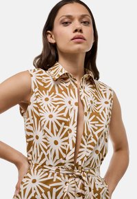 Robe sans manches à boutons en beige avec des motifs floraux blancs, dotée d'un col, d'un fente à l'avant et d'une taille nouée pour une silhouette ajustée.