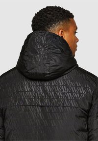 Czarna pikowana kurtka z teksturowanym geometrycznym wzorem, kapturem i gładkim wykończeniem, widziana od tyłu.