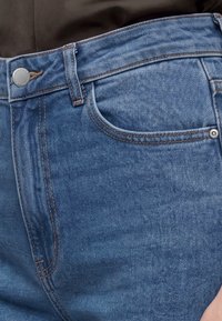 Gros plan de la zone de la taille d'un jean en denim bleu avec des passants de ceinture visibles, une poche avant et un bouton métallique fermé sur une chemise foncée.