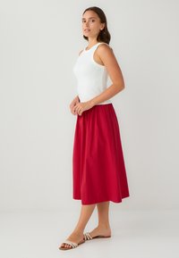 Haut sans manches blanc associé à une jupe midi rouge fluide. La jupe a une texture lisse et une taille élastique. Porté avec des sandales blanches à lanières.