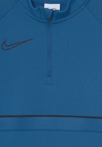 Top deportivo azul con cremallera, fabricado en un tejido suave. Presenta un logo negro de Nike y una franja texturizada horizontal en el centro.