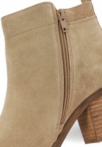 Beige suede ankle boot with a side zipper, textured upper, and a stacked wooden heel. Features smooth stitching and a snug fit.