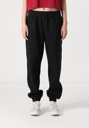 Persona que lleva pantalones deportivos negros sueltos con cintura y puños elásticos, combinados con zapatillas blancas y una camiseta corta roja.