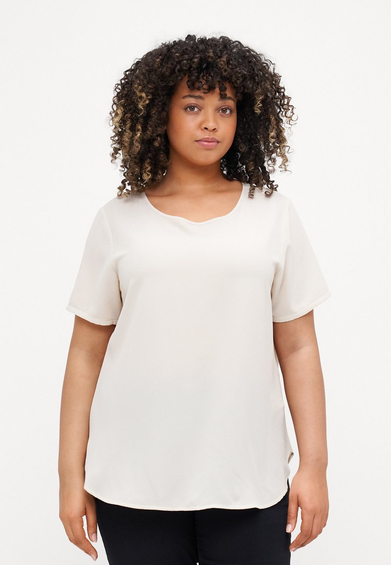 Cream-colored short-sleeve blouse with a scoop neckline, featuring a smooth, lightweight fabric and a slightly curved hem.