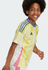 Maglia sportiva gialla e bianca con accenti laterali rosa, caratterizzata da strisce blu sulle maniche e un motivo testurizzato su tutta la superficie.