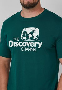 Teal Baumwoll-T-Shirt mit einem weißen Grafikdesign eines Globus und dem Text "THE DISCOVERY CHANNEL", Rundhalsausschnitt, kurze Ärmel, glatte Textur.