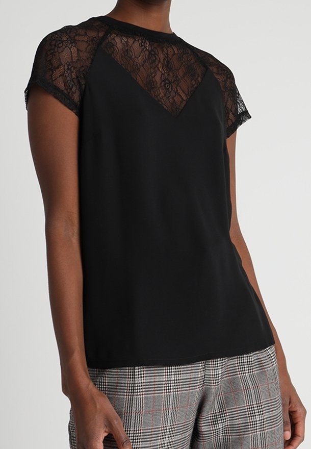 Black short-sleeve top with sheer lace yoke, featuring a round neckline and a relaxed fit. Worn with gray plaid trousers.