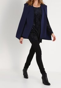 Granatowy płaszcz-kurtka nałożony na fakturowaną czarną sukienkę, połączony z czarnymi legginsami i botkami do kostek. Gładki materiał i dopasowana sylwetka.