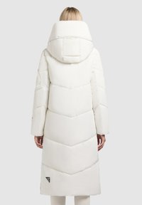 Baltas pūkinis švarkas su zigzago raštu, aukštu apykakle ir logotipo emblema. Turi ilgas rankoves ir vidutinio ilgio dizainą, užtikrinantį šilumą.