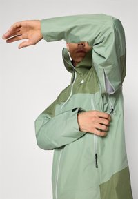 Veste imperméable vert clair avec un col haut, des poignets ajustables et des zips. Présente un design coloré avec des accents verts plus foncés.