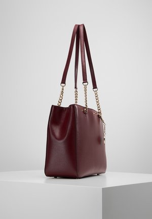 Burgundy leather tote bag with gold chain and leather double shoulder straps, standing on a white pedestal against a neutral background.