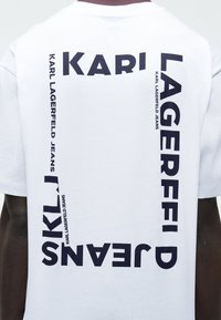 Άσπρο T-shirt με μαύρο έντονο κείμενο «KARL LAGERFELD JEANS», διατεταγμένο σε τετράγωνο στην πλάτη, φορεμένο από άτομο με σκουρόχρωμο δέρμα.