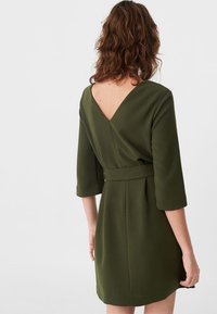 V-päähän ulottuva vihreä mekko, jossa on kolme neljäsosaa hihat, vyötärölle tiivistetty malli vyöllä ja sileä rakenne. Keskivartalon pituinen, hyvin muotoiltu design.