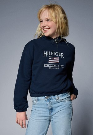 Mornarsko modra dukserica s kapuco, s vezenim napisom "HILFIGER" in oblikom zastave. V kombinaciji z svetlo modrimi kavbojkami z rebrastimi manšeti.