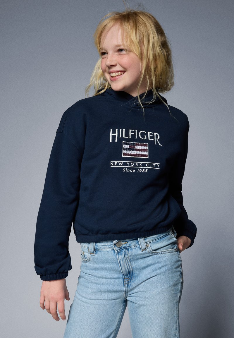 Felpa blu navy con cappuccio, con la scritta ricamata "HILFIGER" e un design a bandiera. Abbinata a jeans azzurri chiari con polsini a listini.