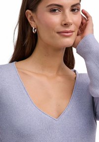 Jasnofioletowy prążkowany sweter z głębokim dekoltem w serek. Długie rękawy o dopasowanym kroju. Modelka ma na sobie srebrne kolczyki koła. Gładka tekstura.