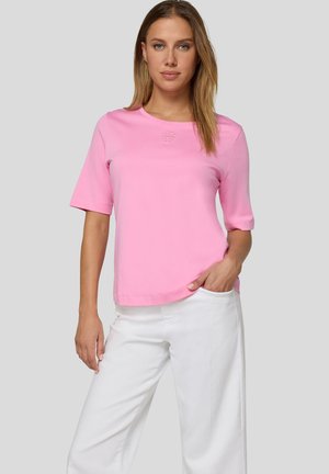Frau trägt ein rosa kurzärmeliges Oberteil und weiße Hose, steht mit einer Hand in der Tasche vor einem schlichten grauen Hintergrund.
