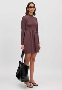 Weinrotes Langarmkleid mit figurbetontem Oberteil und ausgestelltem Rock, kombiniert mit einer schwarzen Tote-Tasche und Flip-Flops. Glatte Textur, schlichtes Design.