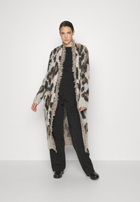 Pletená leopardia srdcová bunda v béžovej a čiernej farbe, s strapcovými okrajmi. Nosená cez čierny top a čierne nohavice, s uvoľneným strihom.
