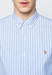 Jasnoniebieska koszula z długim rękawem w pionowe białe paski, z małym haftowanym logo jeźdźca po lewej stronie klatki piersiowej.