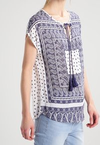 Námořnická a bílá košile s krátkými rukávy, výstřihem do V a třásňovým vázáním. Obsahuje geometrické a květinové vzory po celém povrchu.