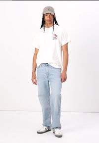 Hvit bomulls t-skjorte med grafisk trykk, lyseblå denimjeans og grå caps. Skoene har svarte og hvite detaljer og en teksturert såle.
