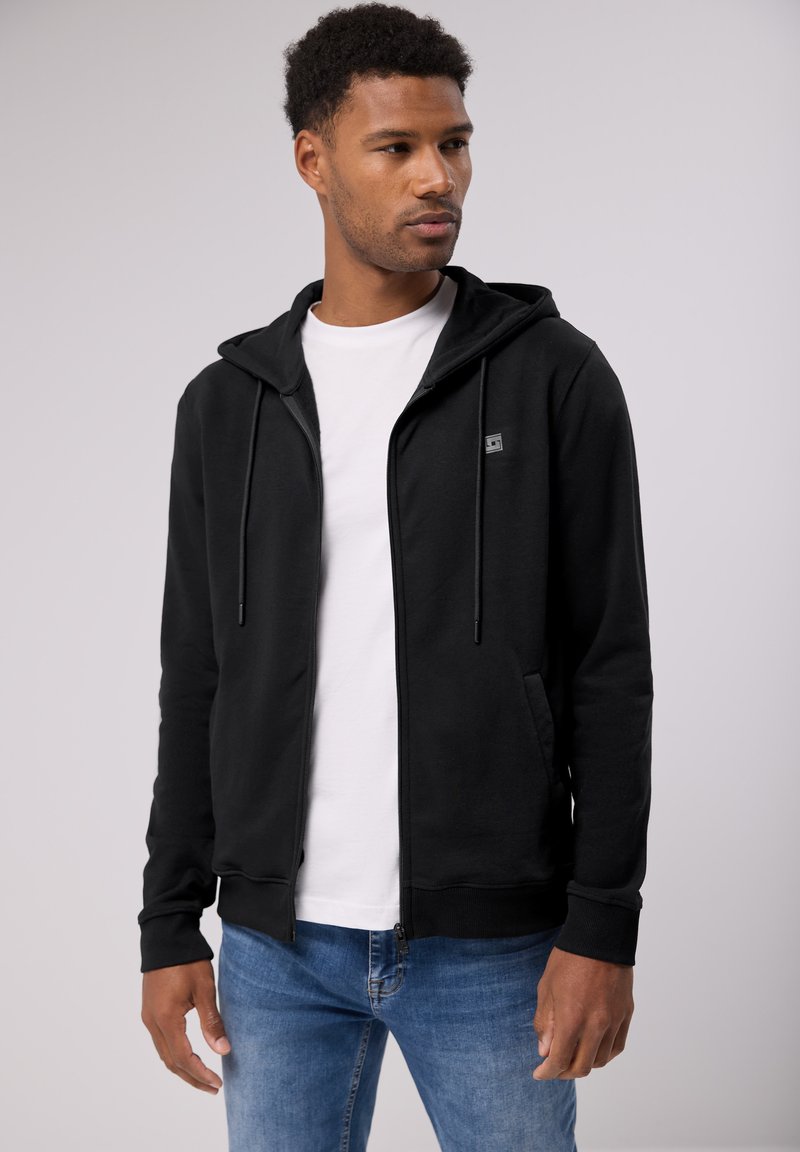 Černá zipová mikina s kapucí na šňůrku, přední kapsy a malé logo na hrudi, kombinovaná s bílým tričkem a modrými džíny.
