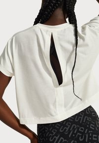 White cropped t-shirt with short sleeves, featuring a back cutout. Fabric appears soft with a smooth texture, worn over black patterned leggings.